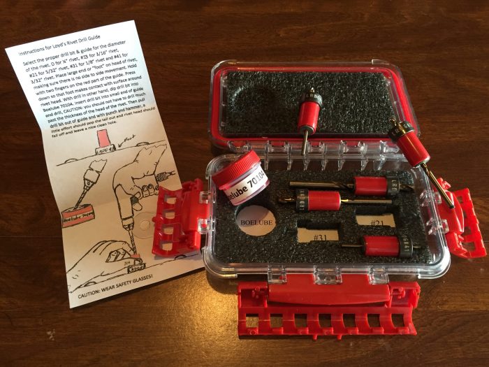 A red plastic case with foam inserts holds various small tools and components. Next to it is an instruction sheet with diagrams. The case is open on a wooden surface.